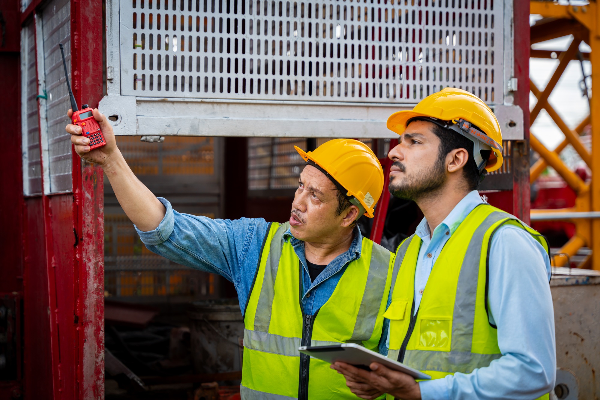 Two men in safety gear are looking at a piece of machinery. They are wearing reflective vests